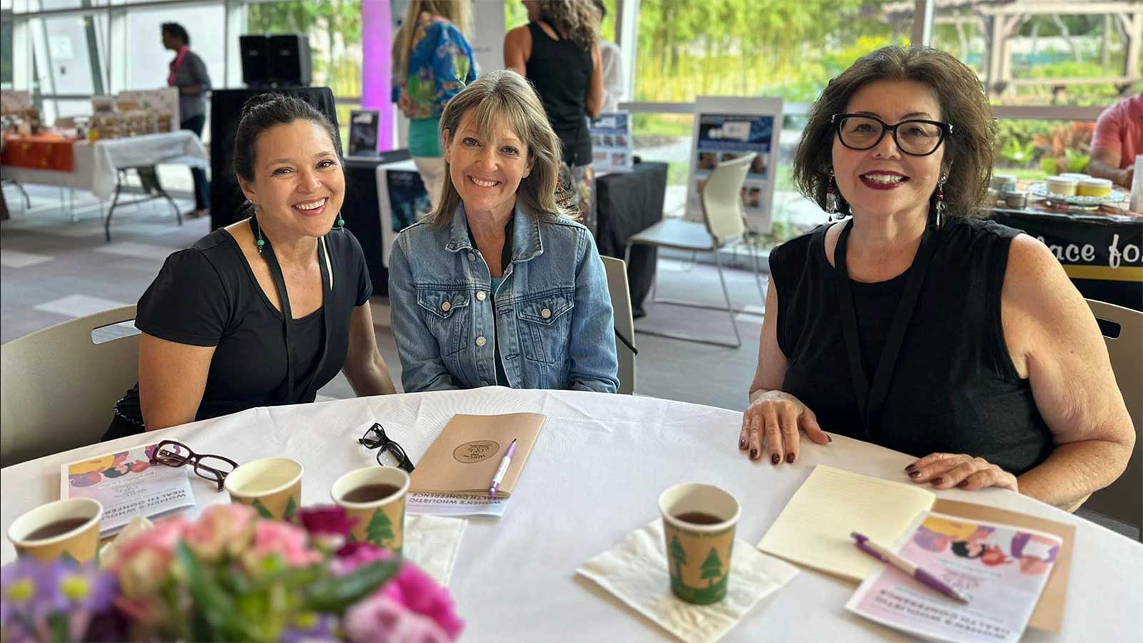 Three smiling women enjoying a corporate wellness event around a table with coffee and brochures, creating a warm, friendly atmosphere.