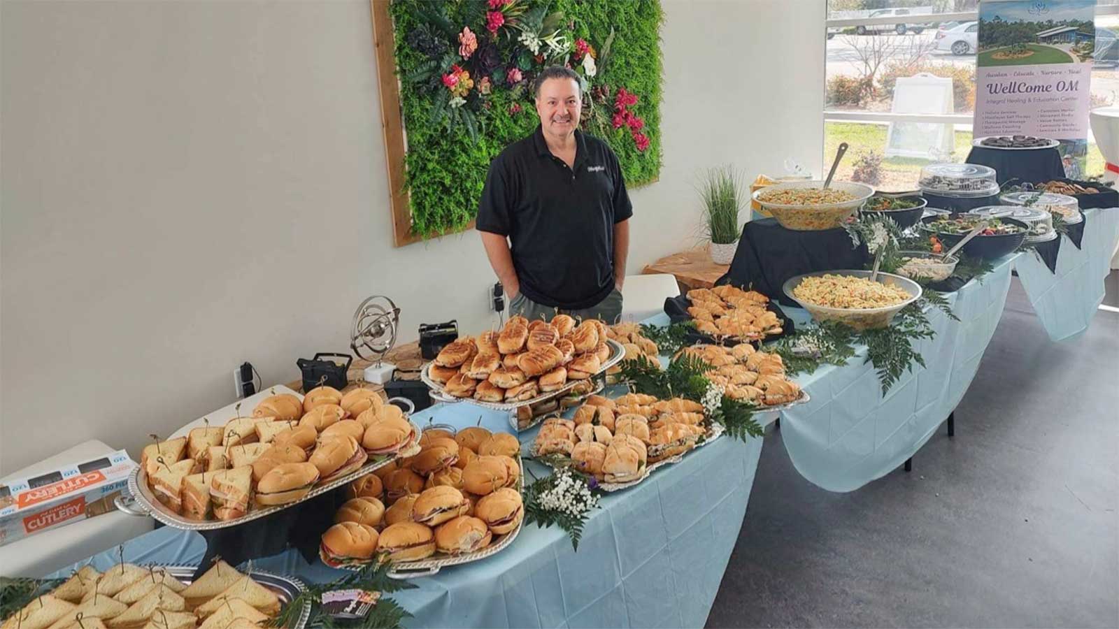 A smiling caterer stands proudly by a well-arranged buffet table filled with a variety of sandwiches and pasta dishes at a Corporate Wellness Event.
